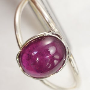 Puede incluir: Anillo de plata con una gran gema ovalada de color magenta oscuro. La gema est&aacute; engastada en un bisel de plata y la banda del anillo es de plata pulida. La gema tiene una superficie lisa y pulida.