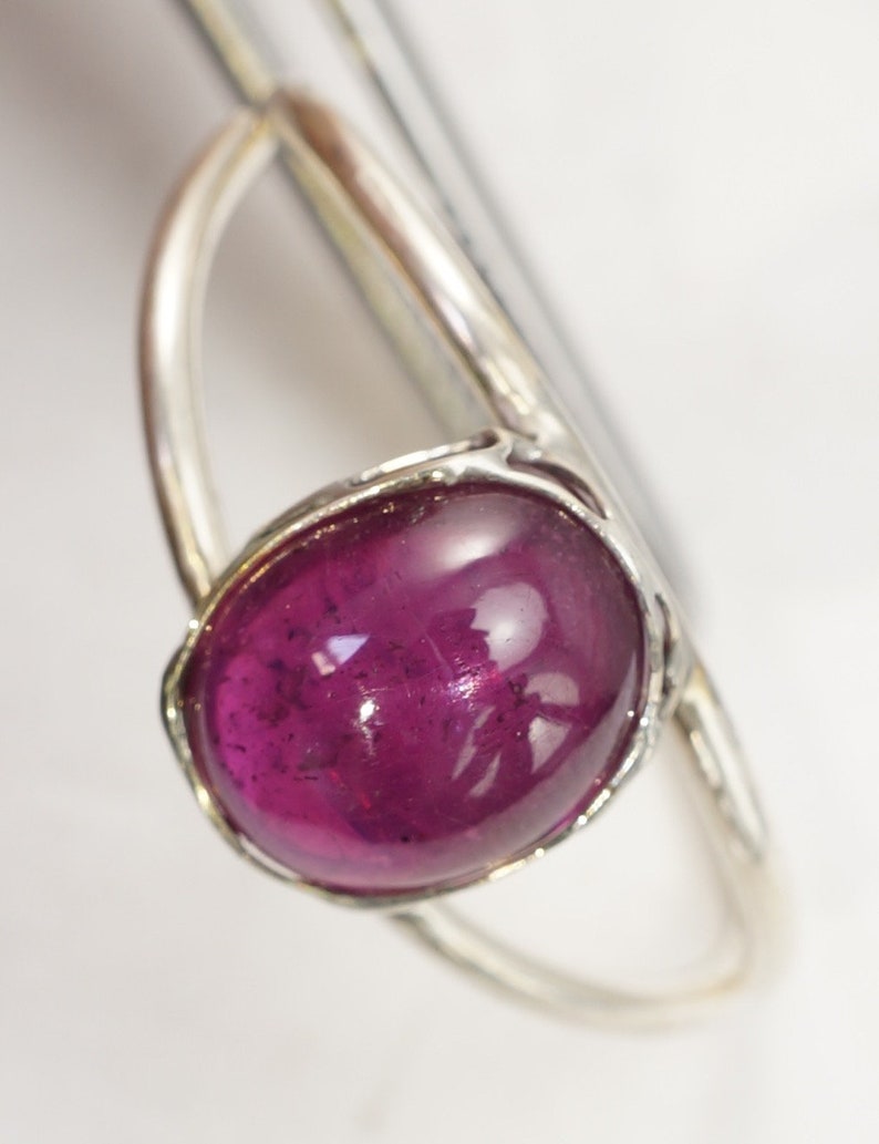 Puede incluir: Anillo de plata con una gran gema ovalada de color magenta oscuro. La gema est&aacute; engastada en un bisel de plata y la banda del anillo es de plata pulida. La gema tiene una superficie lisa y pulida.