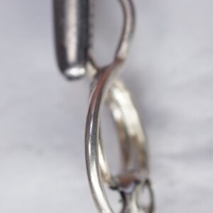 May include: A close-up of a silver pendant with a twisted design. The pendant has a loop at the top and a curved, swirling shape. The metal has a shiny, reflective surface, and the background is a blurred, light color.