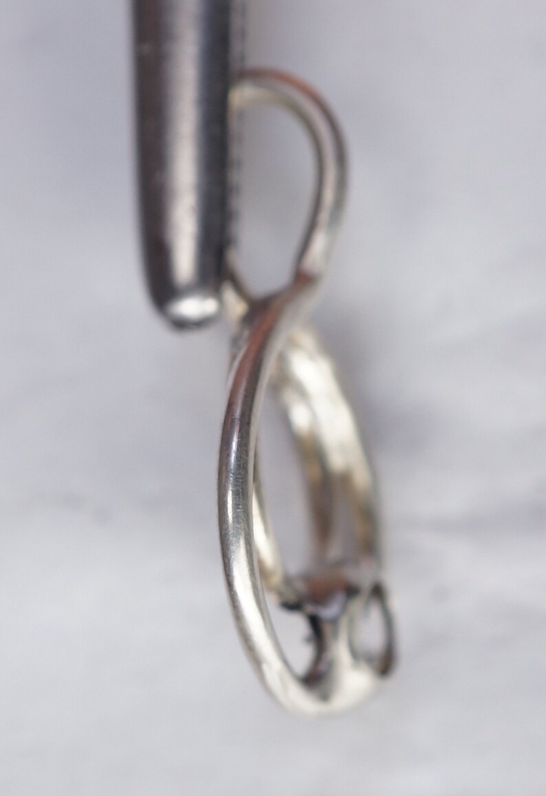 May include: A close-up of a silver pendant with a twisted design. The pendant has a loop at the top and a curved, swirling shape. The metal has a shiny, reflective surface, and the background is a blurred, light color.