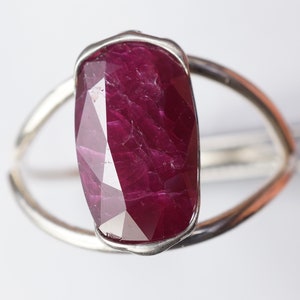 Op de afbeelding: Een zilveren ring met een grote, gefacetteerde, rode robijn edelsteen. De robijn is gezet in een bezel setting en heeft een unieke, onregelmatige vorm.