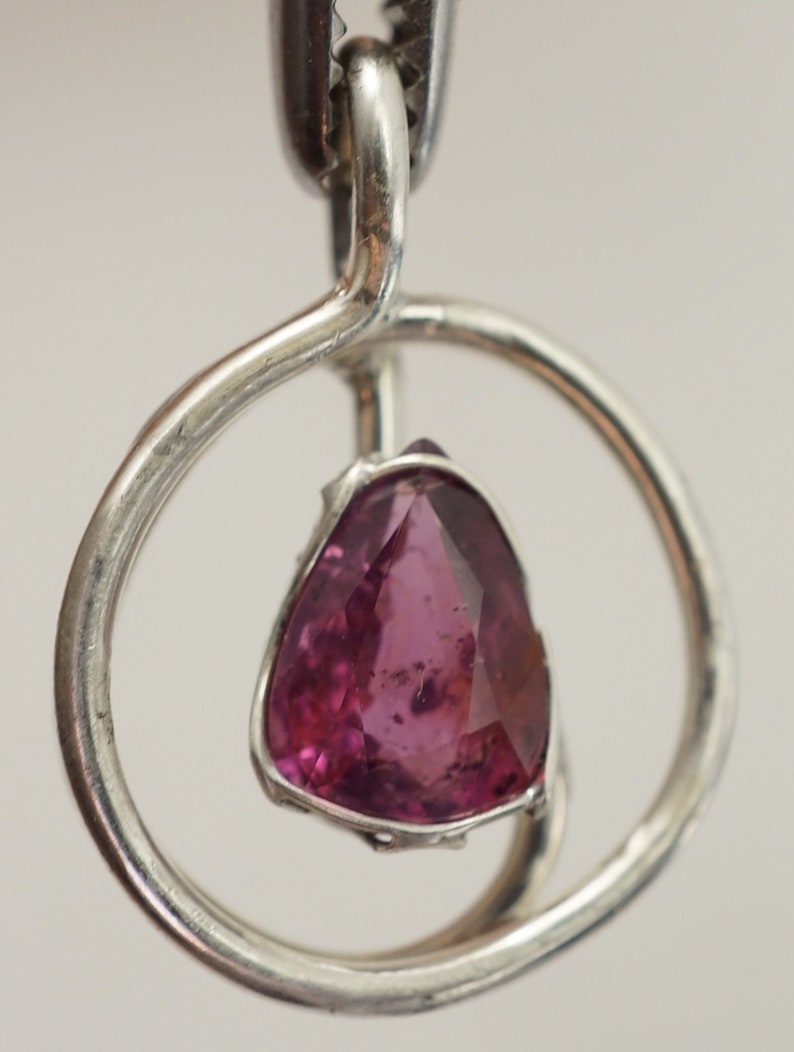 May include: A silver pendant featuring a pink gemstone set within a circular frame. The pendant hangs from a silver bail.