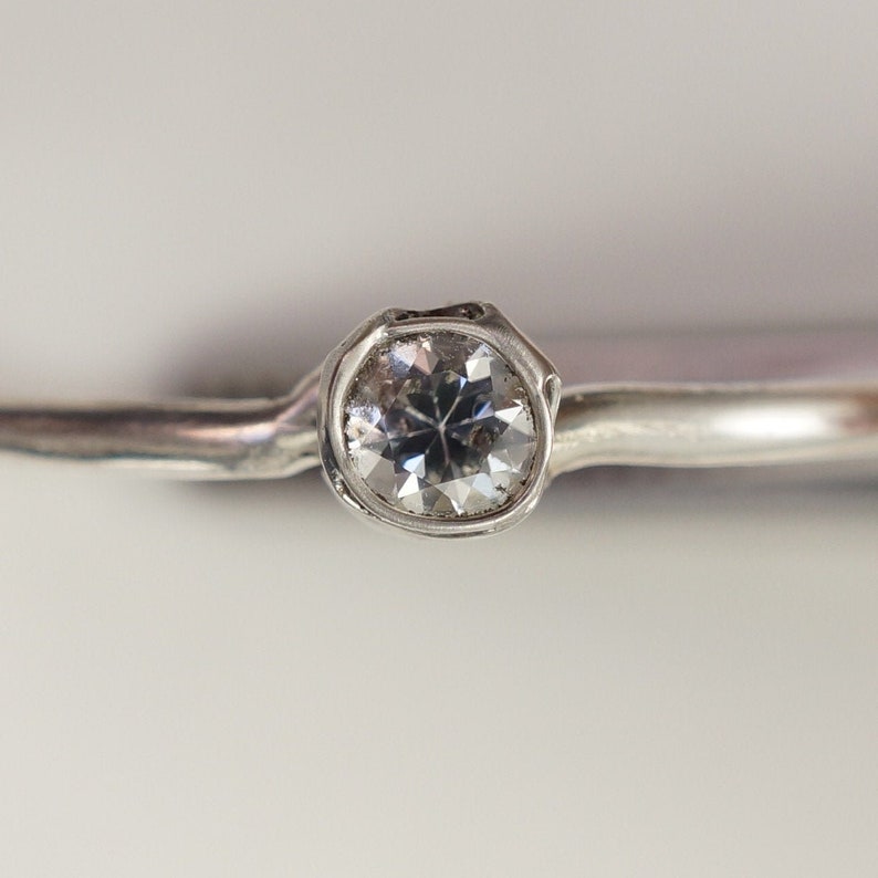 May include: A silver ring with a single, clear, round gemstone set in a textured band.