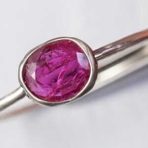 May include: A silver ring featuring a vibrant, oval-cut ruby gemstone. The ruby is a deep pink color and is set in a bezel setting. The ring's band is a simple, polished silver. The ring is a piece of jewelry.