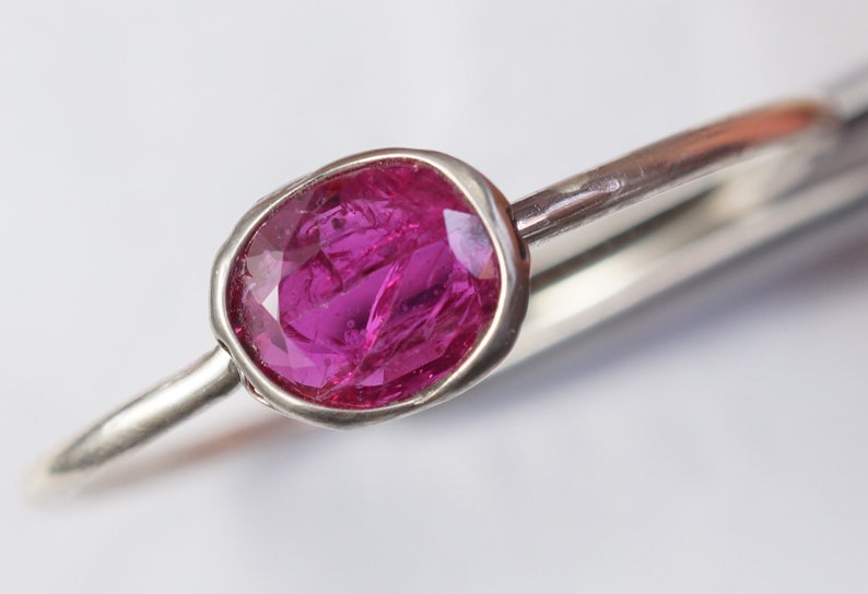 May include: A silver ring featuring a vibrant, oval-cut ruby gemstone. The ruby is a deep pink color and is set in a bezel setting. The ring's band is a simple, polished silver. The ring is a piece of jewelry.