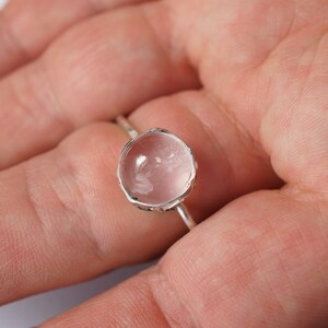 Puede incluir: Un anillo de plata con una piedra preciosa redonda, transparente y facetada. La piedra preciosa tiene un tono rosa claro.