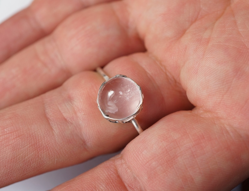 Puede incluir: Un anillo de plata con una piedra preciosa redonda, transparente y facetada. La piedra preciosa tiene un tono rosa claro.