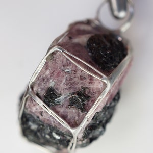 Puede incluir: Un colgante de piedra preciosa rosa y negra con una jaula de alambre de plata. La piedra preciosa es rugosa y tiene una forma natural y org&aacute;nica.