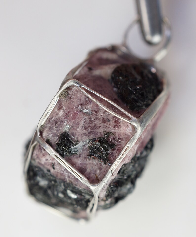 Puede incluir: Un colgante de piedra preciosa rosa y negra con una jaula de alambre de plata. La piedra preciosa es rugosa y tiene una forma natural y org&aacute;nica.