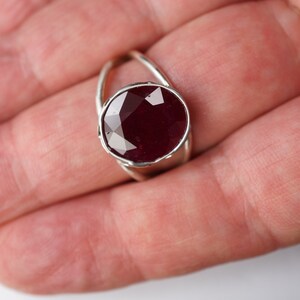 May include: A silver ring with a large, faceted ruby gemstone set in a bezel setting. The ring has a delicate, twisted band.