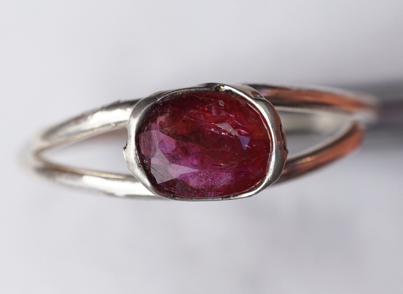 Puede incluir: Un anillo de plata con una piedra ovalada de color rosa oscuro engastada en un engaste texturizado.