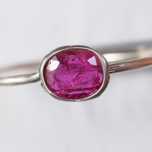May include: A silver ring featuring a vibrant, oval-cut ruby gemstone. The ruby is a deep magenta color, set in a bezel setting. The band is thin and polished, creating a minimalist design. This ring is a piece of jewelry.