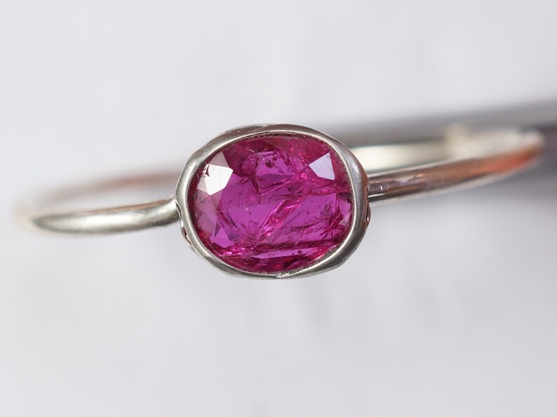 May include: A silver ring featuring a vibrant, oval-cut ruby gemstone. The ruby is a deep magenta color, set in a bezel setting. The band is thin and polished, creating a minimalist design. This ring is a piece of jewelry.
