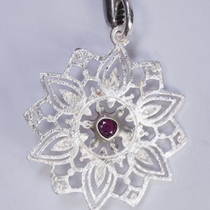 May include: A silver pendant featuring a delicate, intricate design resembling a flower. The centre of the pendant features a small, dark purple gemstone.