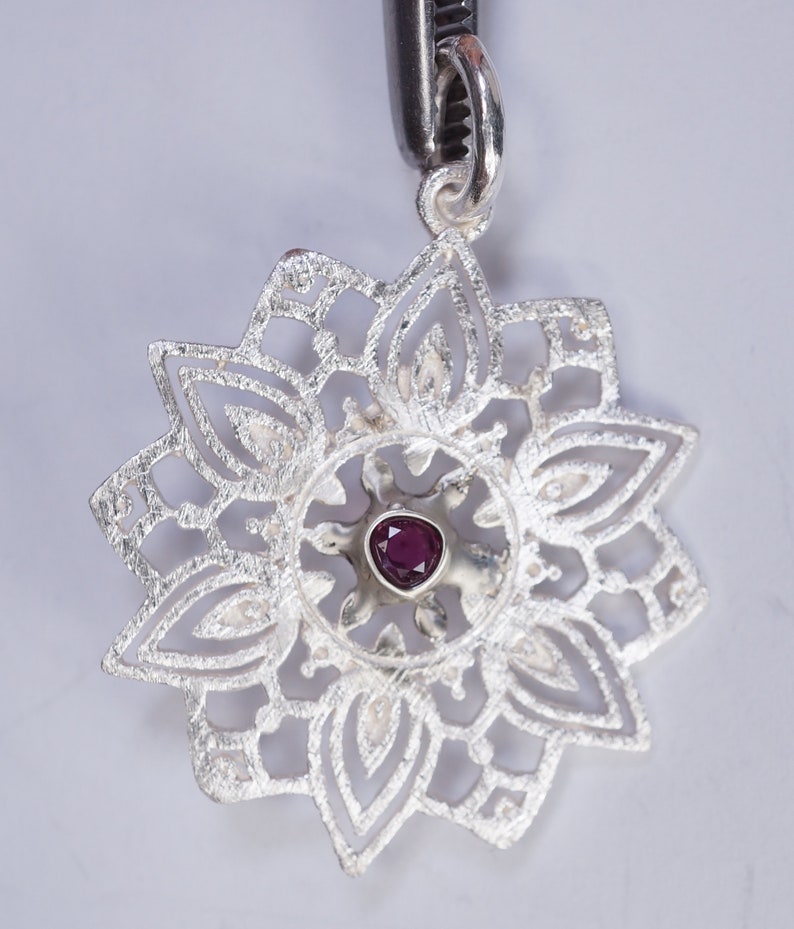May include: A silver pendant featuring a delicate, intricate design resembling a flower. The centre of the pendant features a small, dark purple gemstone.