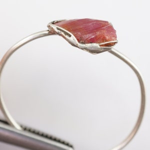 May include: A silver ring featuring a raw pink ruby gemstone set in a delicate wire cage.