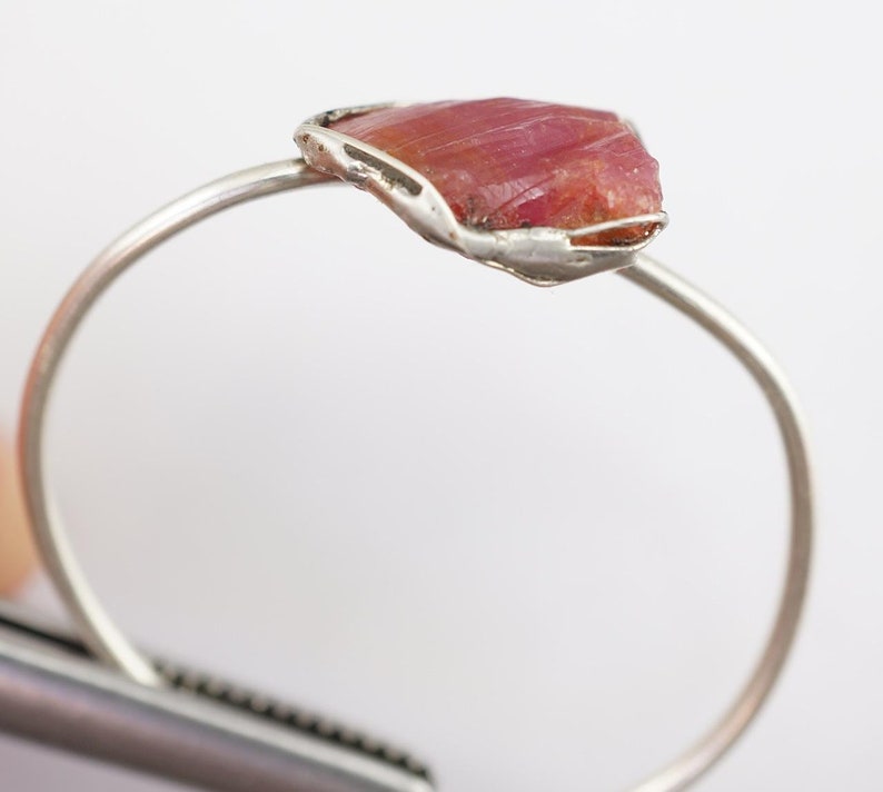 May include: A silver ring featuring a raw pink ruby gemstone set in a delicate wire cage.