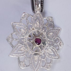 May include: A silver pendant with a delicate, intricate design featuring a central ruby gemstone. The pendant is shaped like a flower with multiple layers of petals.