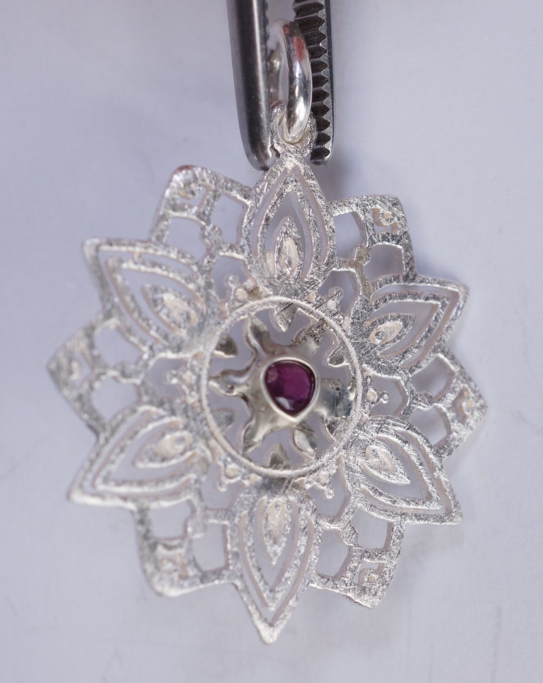 May include: A silver pendant with a delicate, intricate design featuring a central ruby gemstone. The pendant is shaped like a flower with multiple layers of petals.