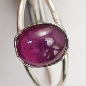 Puede incluir: Un anillo de plata con una gema grande, ovalada y de color magenta intenso. La gema est&aacute; engastada en un bisel y tiene una superficie pulida y reflectante. El anillo tiene un dise&ntilde;o abierto &uacute;nico.