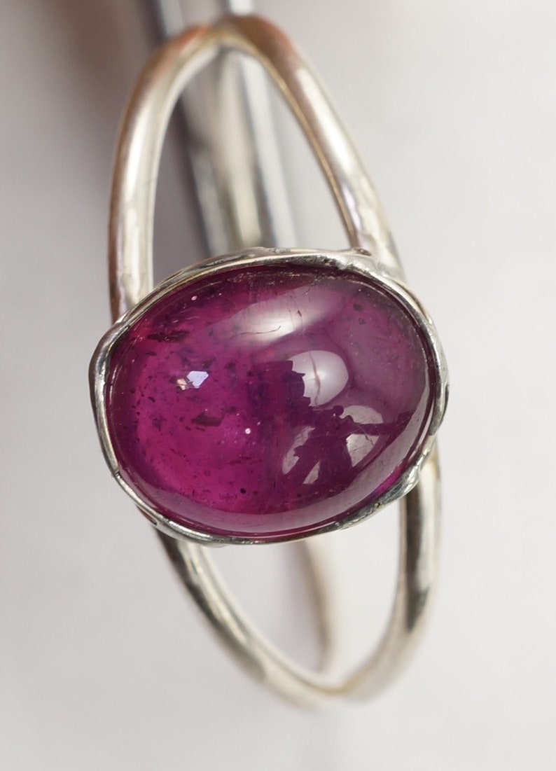 Puede incluir: Un anillo de plata con una gema grande, ovalada y de color magenta intenso. La gema est&aacute; engastada en un bisel y tiene una superficie pulida y reflectante. El anillo tiene un dise&ntilde;o abierto &uacute;nico.