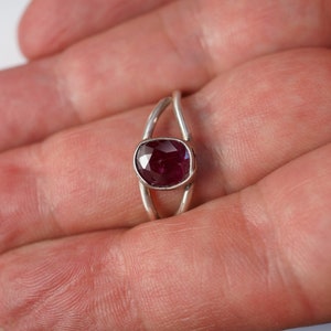 K&ouml;nnte beinhalten: Ein silberner Ring mit einem tief roten ovalen Edelstein, der in der Mitte eingesetzt ist. Der Ring hat ein einzigartiges Design mit zwei B&auml;ndern, die sich &uuml;berkreuzen.