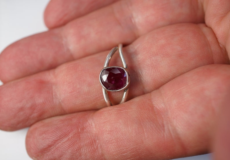 K&ouml;nnte beinhalten: Ein silberner Ring mit einem tief roten ovalen Edelstein, der in der Mitte eingesetzt ist. Der Ring hat ein einzigartiges Design mit zwei B&auml;ndern, die sich &uuml;berkreuzen.