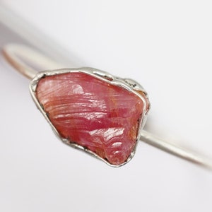 May include: A silver ring featuring a raw pink ruby gemstone set in a wire cage. The ruby has a rough, natural texture and a deep pink colour.