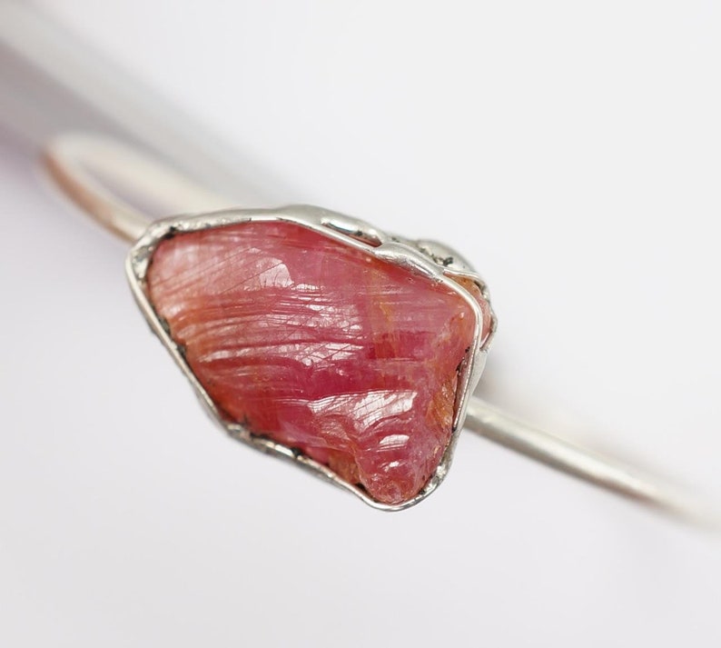 May include: A silver ring featuring a raw pink ruby gemstone set in a wire cage. The ruby has a rough, natural texture and a deep pink colour.