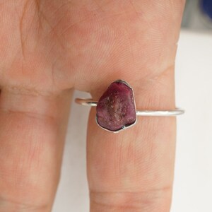 May include: A silver ring with a raw, pink ruby gemstone. The ring is on a finger.
