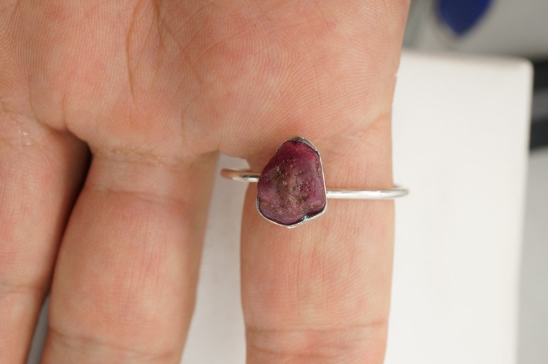 May include: A silver ring with a raw, pink ruby gemstone. The ring is on a finger.