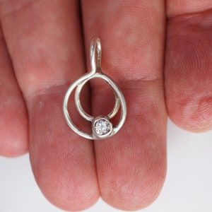 May include: A silver pendant with a small, clear gemstone set in the center. The pendant has a unique design with two interconnected circular shapes. The bail is a simple loop for a chain.