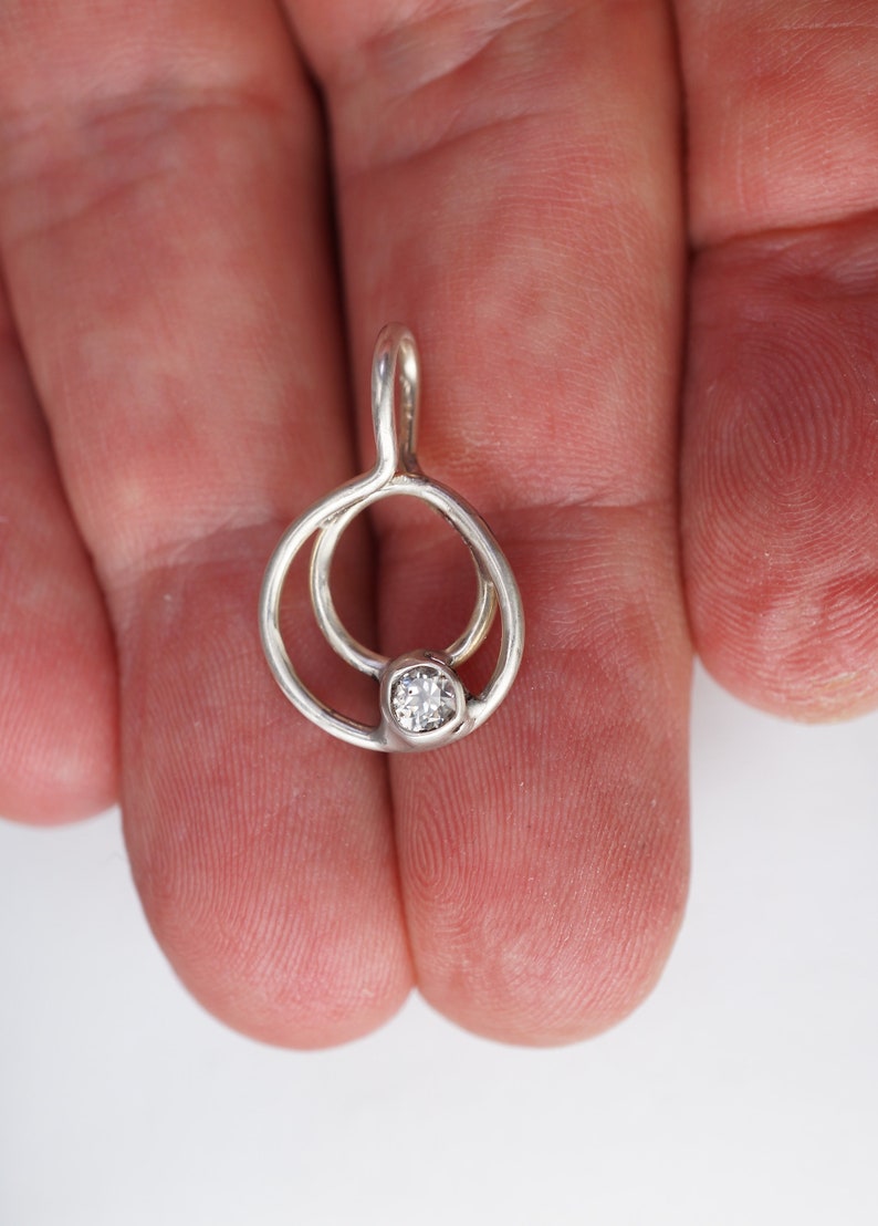 May include: A silver pendant with a small, clear gemstone set in the center. The pendant has a unique design with two interconnected circular shapes. The bail is a simple loop for a chain.