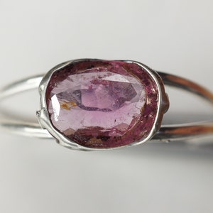 K&ouml;nnte beinhalten: Ein silberner Ring mit einem rosafarbenen ovalen Edelstein, der in einer strukturierten Silberfassung sitzt.