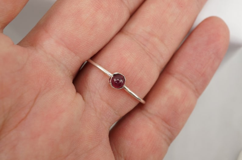 K&ouml;nnte beinhalten: Ein silberner Ring mit einem kleinen, runden, dunkelroten Edelstein.