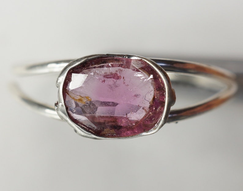 K&ouml;nnte beinhalten: Ein silberner Ring mit einem ovalen rosafarbenen Edelstein, der in einer Zargenfassung gefasst ist. Der Ring hat ein schlichtes, minimalistisches Design.