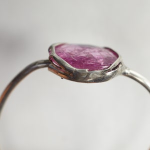 May include: A silver ring with a pink gemstone set in a rough, irregular shape. The ring is made of a thin band of metal and the gemstone is set in a bezel setting.