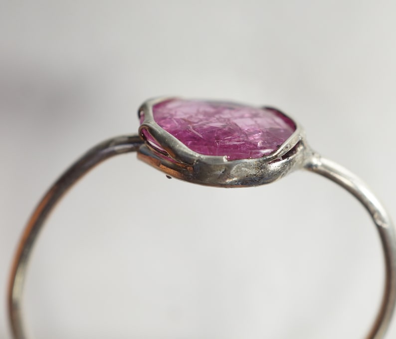 May include: A silver ring with a pink gemstone set in a rough, irregular shape. The ring is made of a thin band of metal and the gemstone is set in a bezel setting.