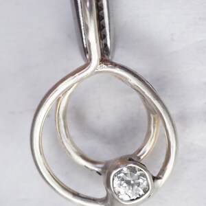 May include: A silver ring with a clear, square-cut gemstone. The ring features a unique design with two interlocking loops and a bezel setting for the gemstone. The ring is held by a pair of silver pliers.