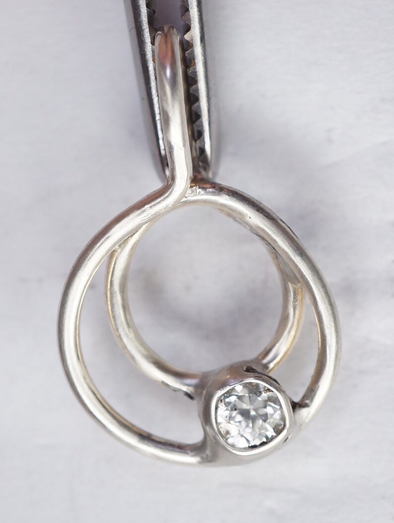 May include: A silver ring with a clear, square-cut gemstone. The ring features a unique design with two interlocking loops and a bezel setting for the gemstone. The ring is held by a pair of silver pliers.