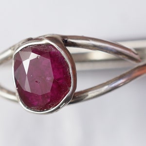 K&ouml;nnte beinhalten: Ein silberner Ring mit einem tief roten Rubin-Edelstein, der in einer Zargenfassung gefasst ist. Der Ring hat ein einzigartiges, verdrehtes Band.
