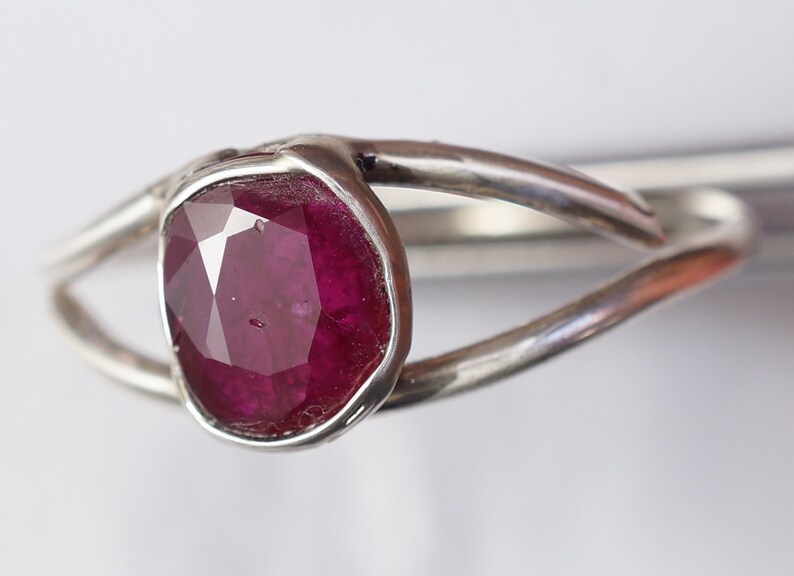 K&ouml;nnte beinhalten: Ein silberner Ring mit einem tief roten Rubin-Edelstein, der in einer Zargenfassung gefasst ist. Der Ring hat ein einzigartiges, verdrehtes Band.