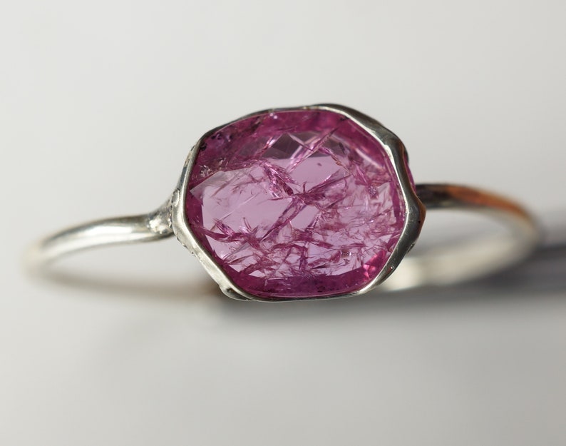 May include: A silver ring with a large, faceted, pink gemstone. The gemstone has a unique, natural, crystalline structure.