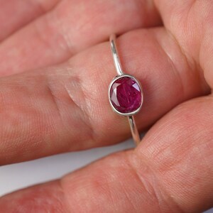 May include: A silver ring featuring a vibrant, oval-cut ruby gemstone. The ruby is a deep, rich red color and is set in a simple, elegant band. The ring is shown on a hand, highlighting the gemstone's brilliance.