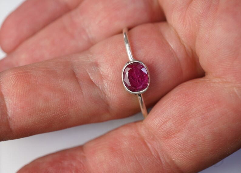 May include: A silver ring featuring a vibrant, oval-cut ruby gemstone. The ruby is a deep, rich red color and is set in a simple, elegant band. The ring is shown on a hand, highlighting the gemstone's brilliance.