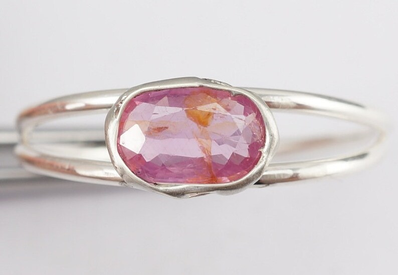 Puede incluir: Un anillo de plata con una piedra ovalada rosa engastada en un engaste de bisel. El anillo tiene una banda doble.