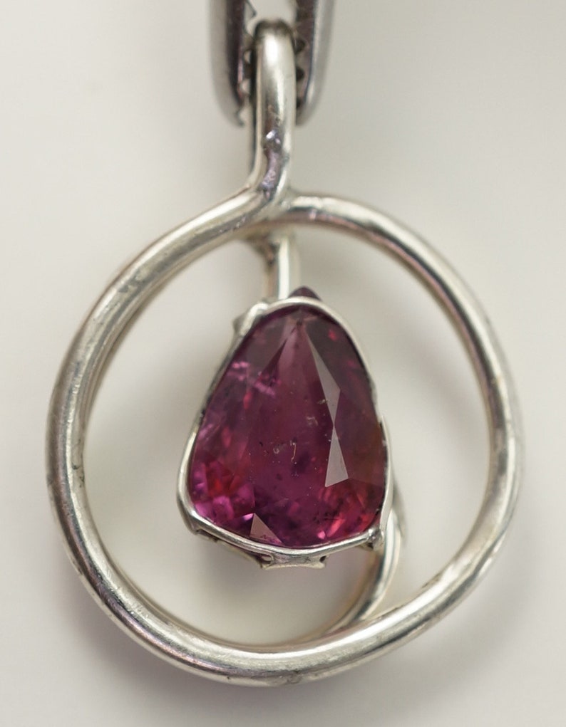 May include: A silver pendant featuring a pink gemstone set within a circular design.