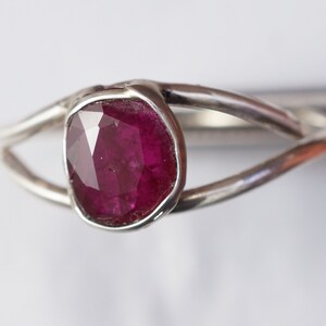 K&ouml;nnte beinhalten: Ein silberner Ring mit einem tiefrosa Edelstein, der in einer Zargenfassung gefasst ist. Der Ring hat ein schlichtes, modernes Design.