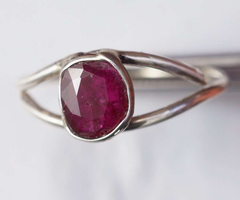 K&ouml;nnte beinhalten: Ein silberner Ring mit einem tiefrosa Edelstein, der in einer Zargenfassung gefasst ist. Der Ring hat ein schlichtes, modernes Design.