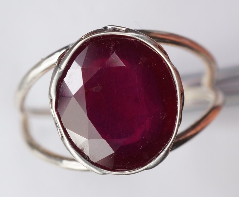 May include: A silver ring with a large, faceted ruby gemstone. The ruby is a deep red color and has a slightly oval shape.